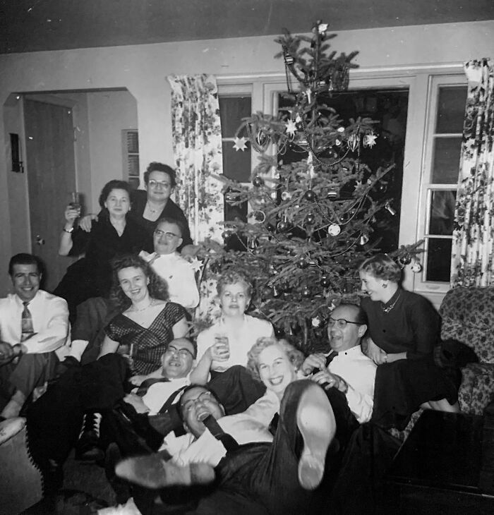 Group of people share found photos celebrating by a decorated Christmas tree in a cozy living room setting.
