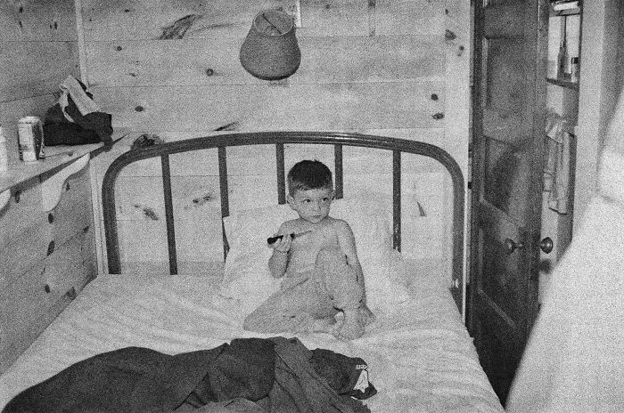 Black and white photo of a young boy sitting on a bed in a rustic wooden room, a found photo shared by people.