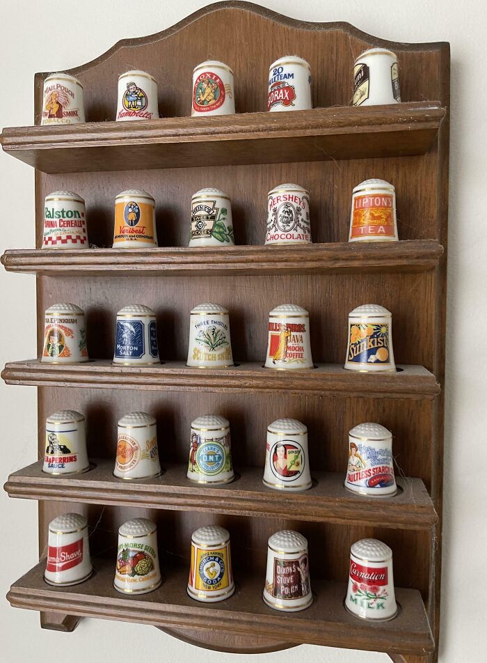 Wooden display shelf holding a collection of vintage advertising ceramic thimbles in a home setting.