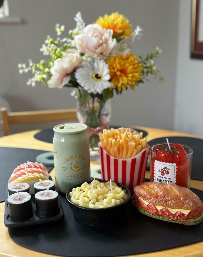 Unusual food-themed candle collection including sushi, fries, macaroni, sandwich, and soda can on a table.