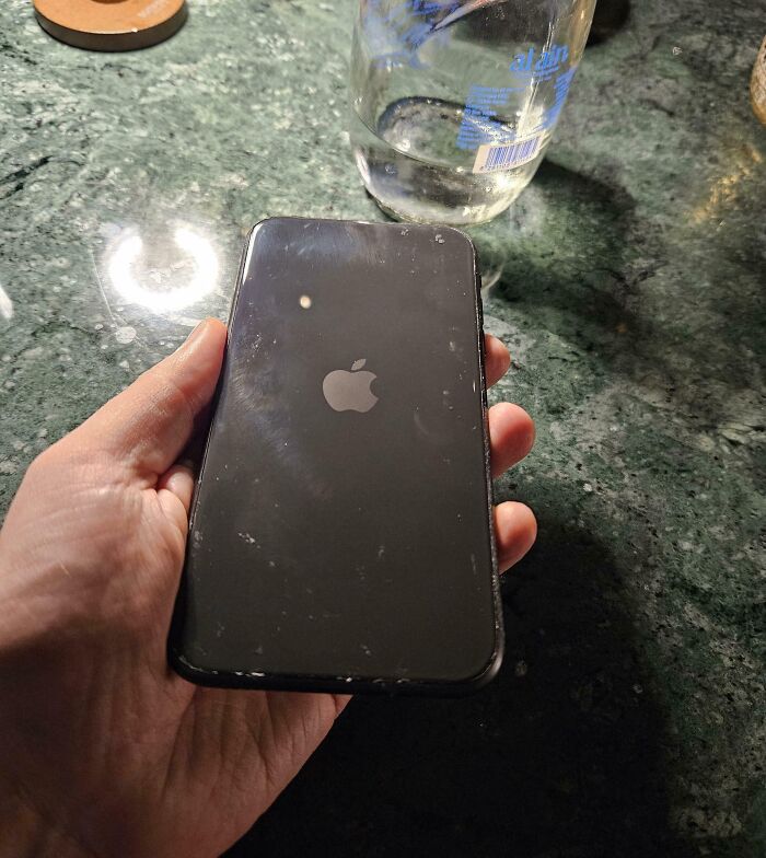 Hand holding a scratched iPhone on a green marble surface next to a glass jug, for curious minds learning about the world.