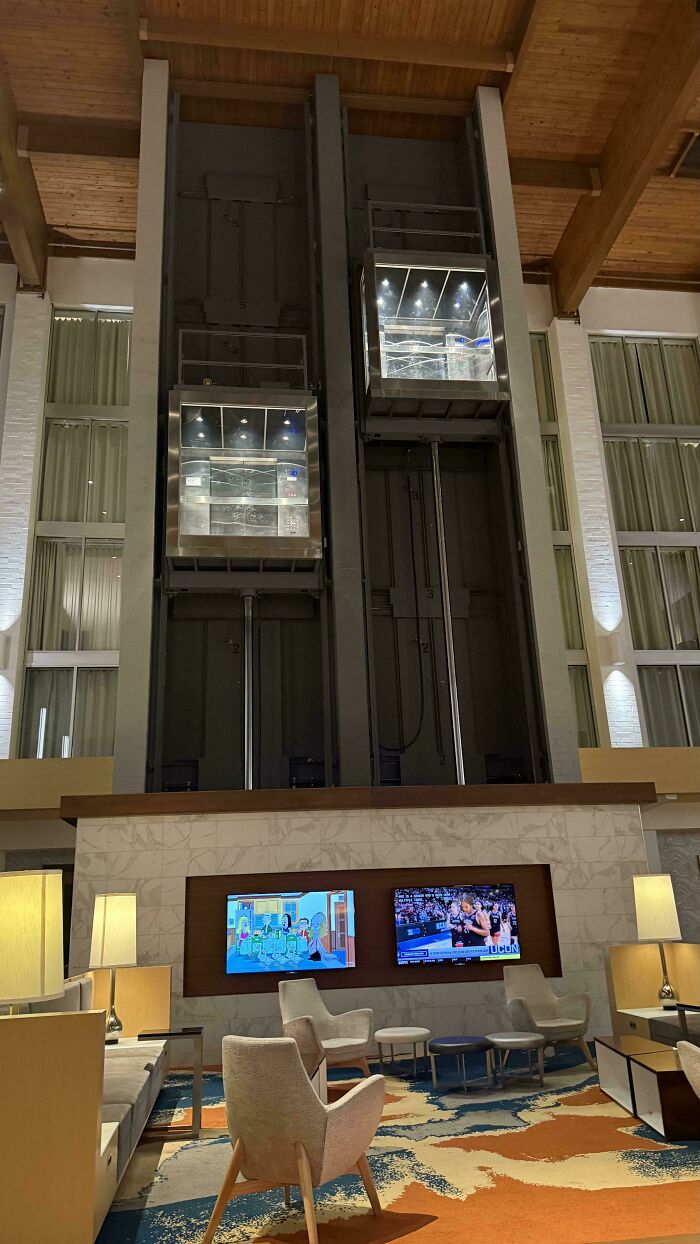 Modern hotel lobby featuring genius design decisions with glass elevators and stylish seating area with TVs and warm lighting.