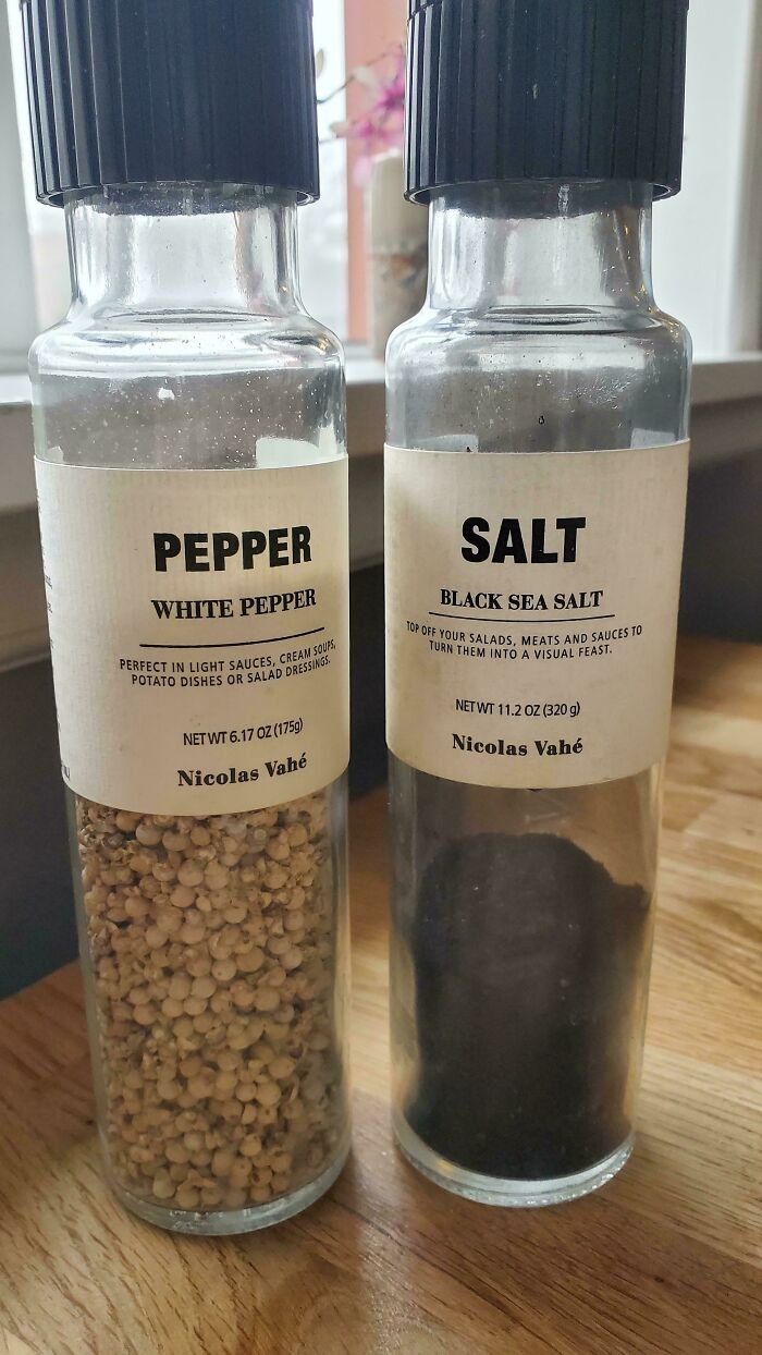 Two clear glass spice grinders with black tops containing white pepper and black sea salt on a wooden surface for hotel design decisions.