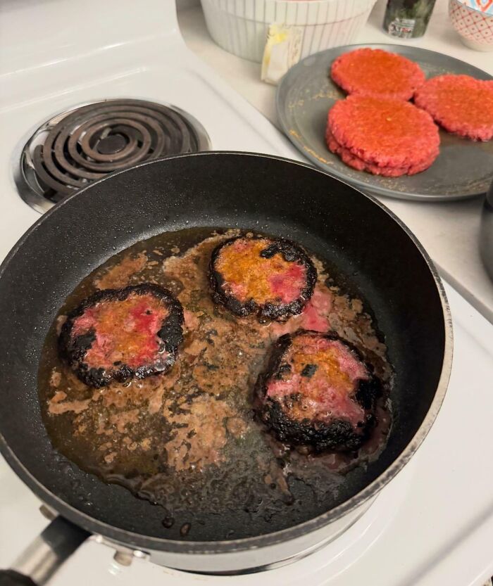 Burnt burger patties cooking in a black pan with raw patties on a plate nearby, showcasing funny cooking fails.