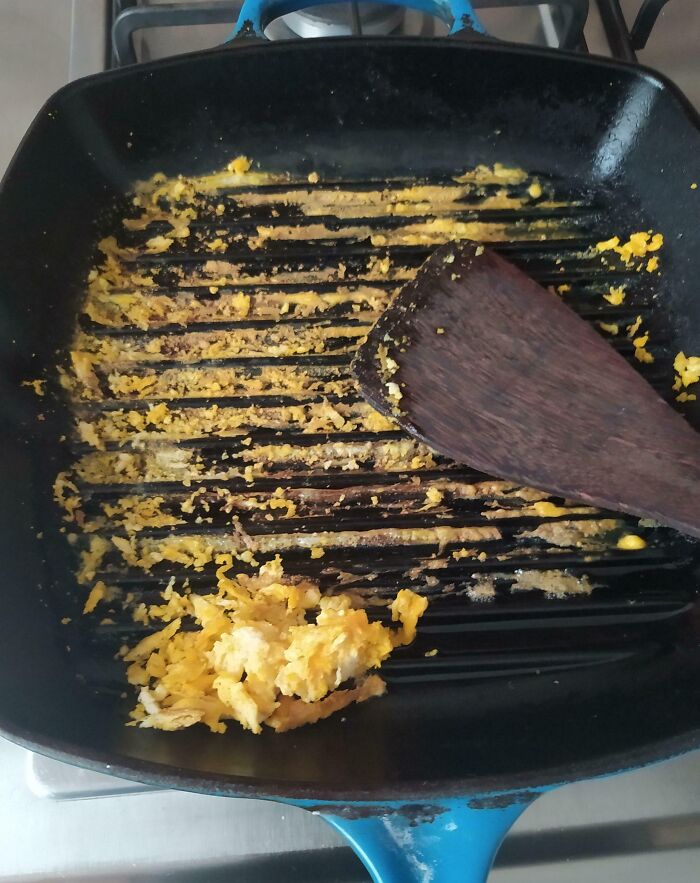 Burnt scrambled eggs stuck to a grill pan with a wooden spatula, showcasing a cooking fail accident in the kitchen.