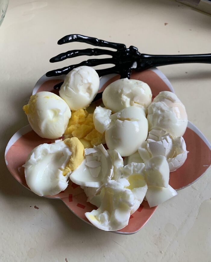 Poorly peeled hard-boiled eggs with broken shells on a plate next to a black skeletal hand utensil, cooking fails.