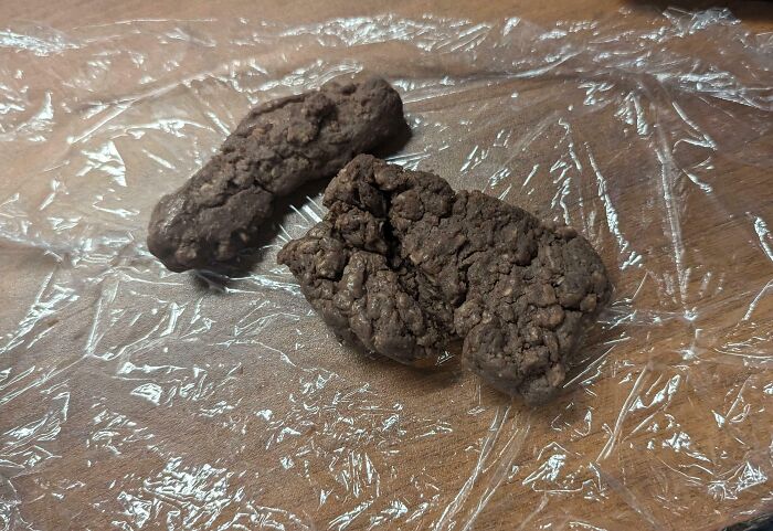 Two poorly baked, misshapen chocolate cookies on plastic wrap showing cooking fails and accidents.