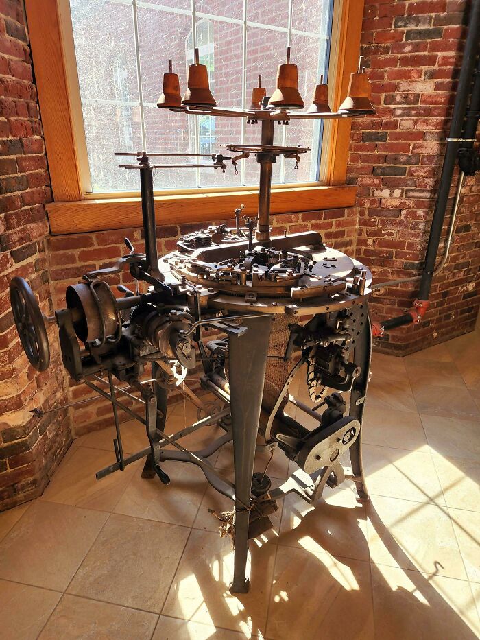 Antique industrial knitting machine displayed in a sunlit room with brick walls, showing how things used to look in textile production.