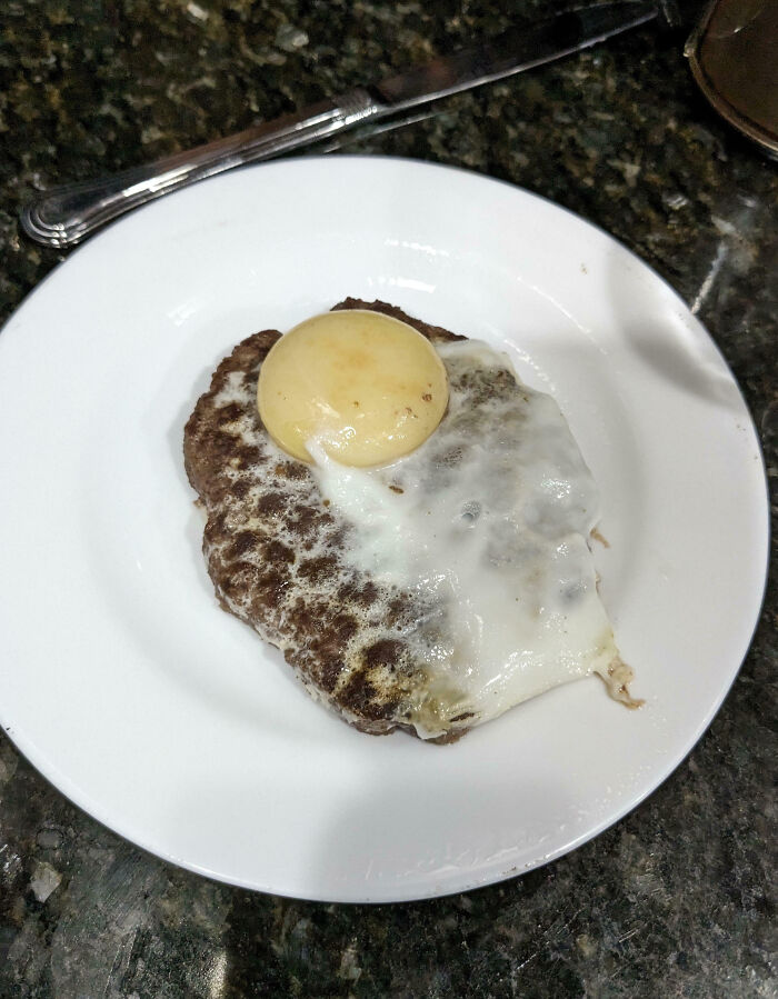 Burnt burger patty with partially cooked egg on top on a white plate, showcasing a cooking fail and accident.