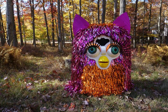 Colorful DIY project resembling a creature with large eyes and tinsel fur, set outdoors in an autumn forest scene.