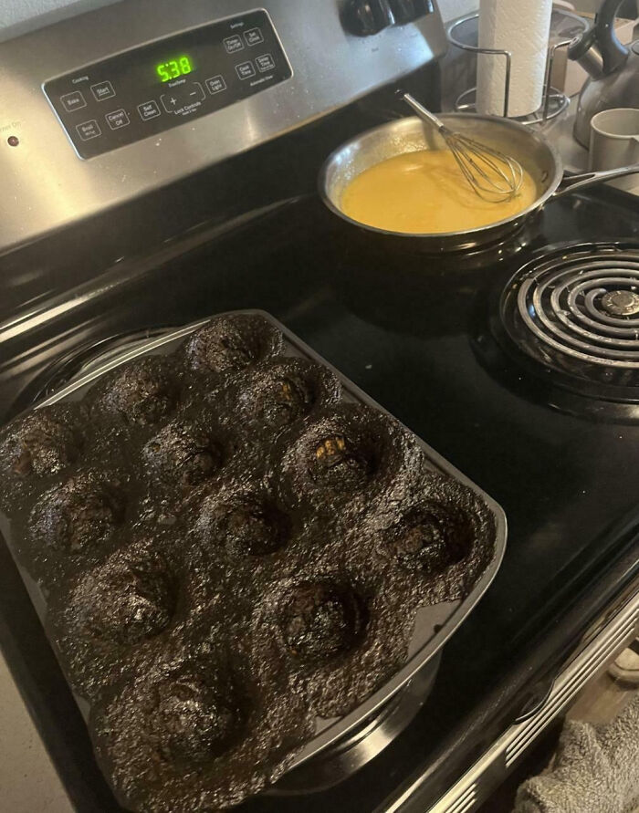 Burnt muffins in a muffin tin on stove next to pan with batter, illustrating cooking fails and accidents.