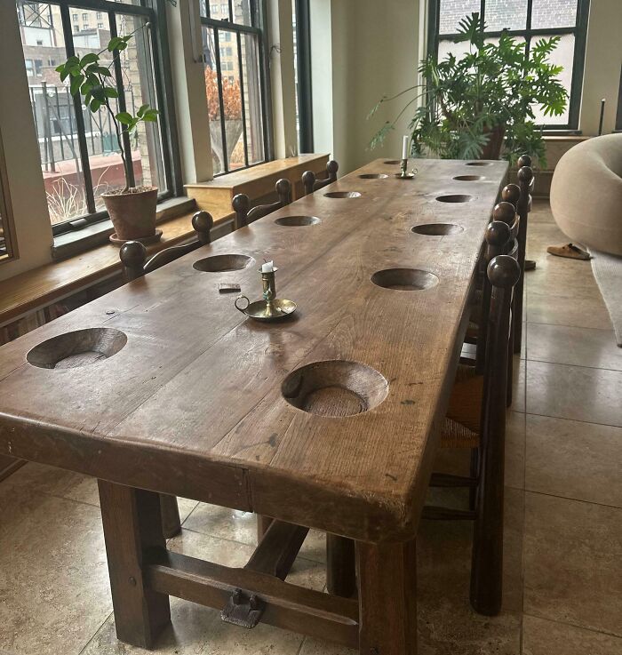 Long antique wooden dining table with carved circular indentations and vintage candlesticks in a bright room with plants.