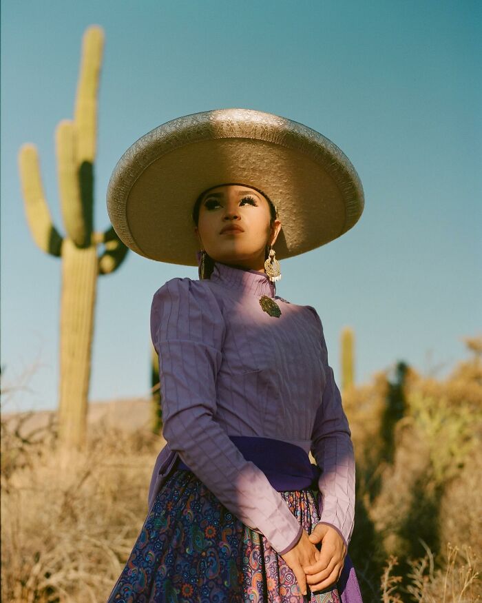 Female rodeo rider in traditional attire and wide-brimmed hat posing confidently in a desert landscape with cacti.