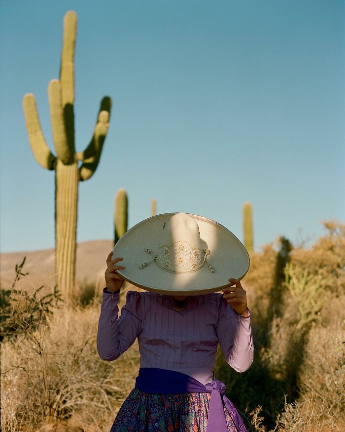 Female rodeo rider holding a traditional hat in a desert landscape with tall cacti, showcasing beauty and power.