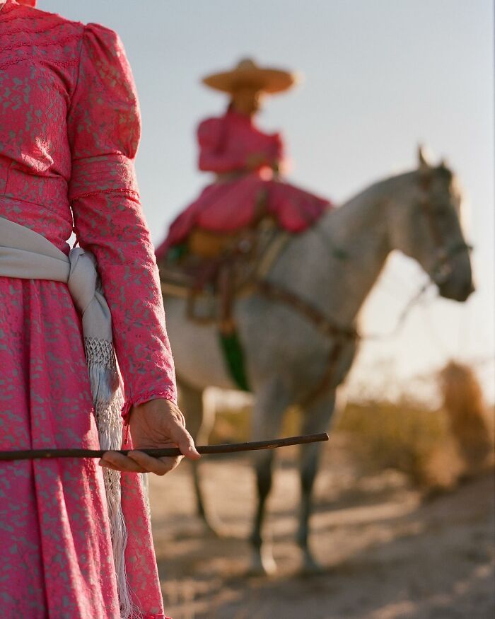 Female rodeo riders dressed in traditional attire with one holding a whip and another riding a horse in the background.