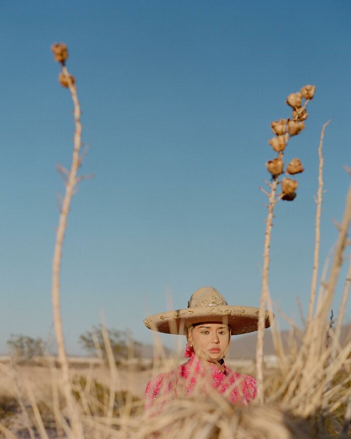 Female rodeo rider wearing a traditional hat and pink outfit standing outdoors in a desert landscape with clear blue sky.