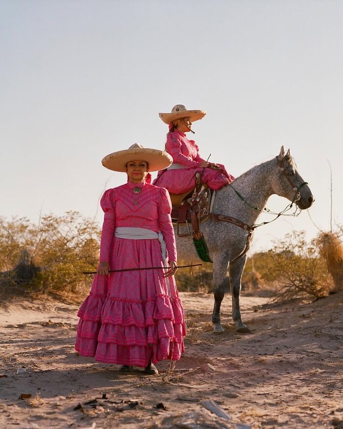 Two all-female rodeo riders in traditional pink dresses and wide-brimmed hats, one standing and one on a gray horse.