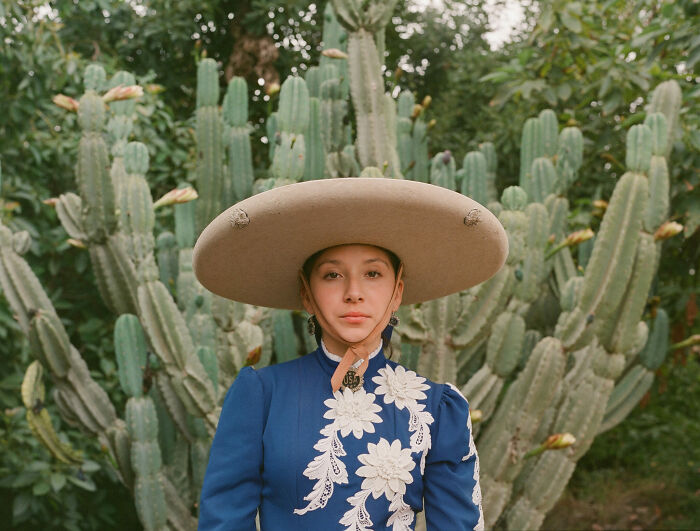 Female rodeo rider wearing traditional outfit and wide-brimmed hat, standing confidently in front of large cactus plants.