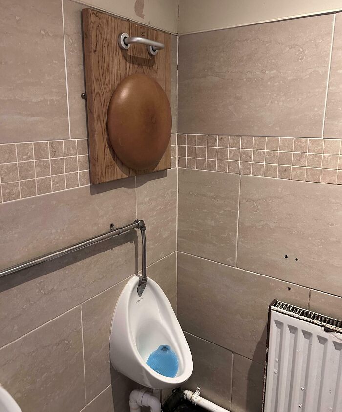 Wall-mounted wooden board with a frying pan lid above a urinal, a clever solution to an overlooked issue in a restroom.