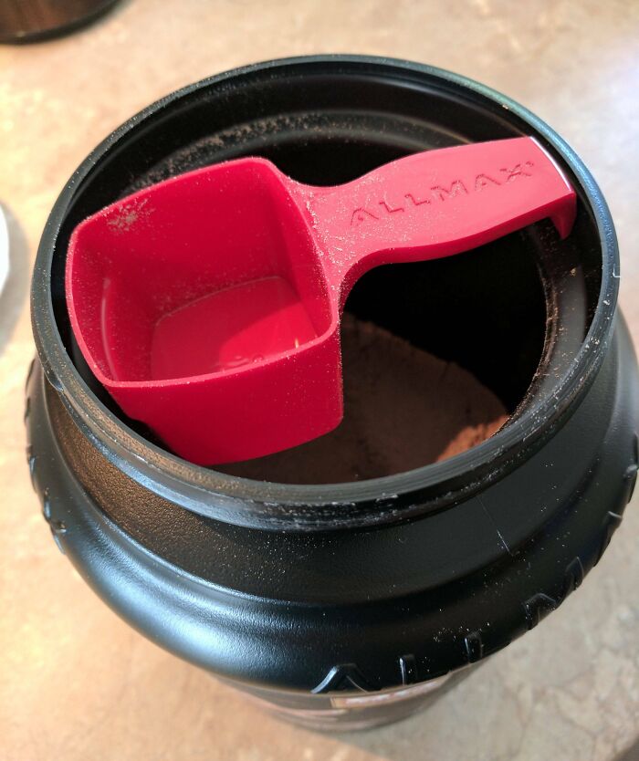 Red measuring scoop inserted in protein powder container showing clever solution for overlooked storage issues.