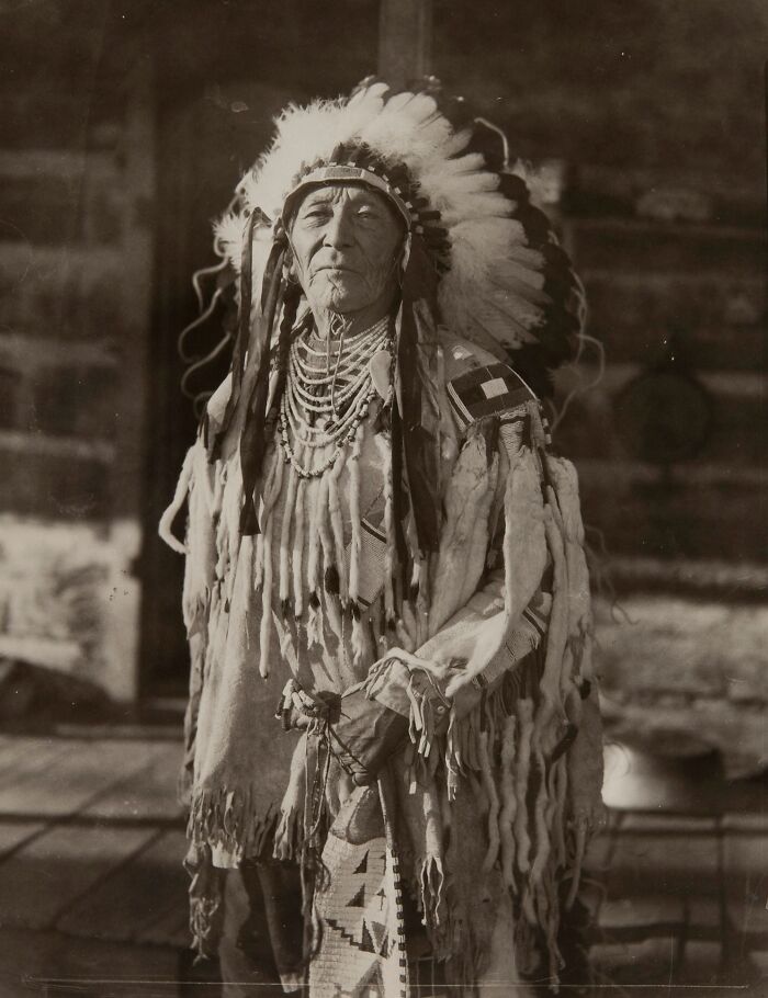 Native American man in traditional dress and feathered headdress in a powerful portrait from over a century ago.