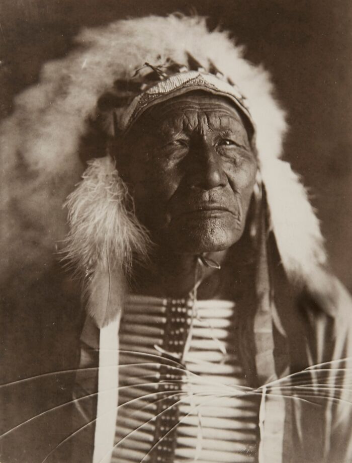 Elder Native American man in traditional headdress and clothing in a powerful historic portrait from over a century ago.