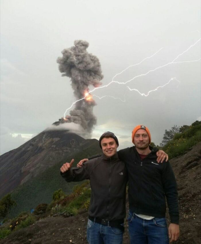 Two men posing near erupting volcano with lightning strikes, showcasing rare natural phenomena in a dramatic landscape.
