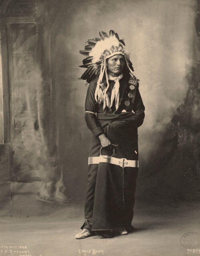 Native American man wearing feathered headdress and traditional attire in a powerful portrait from over a century ago.