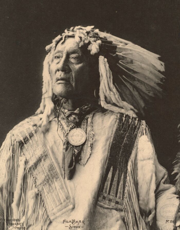 Native American man in traditional Sioux attire and feathered headdress in a powerful historical portrait from over a century ago.