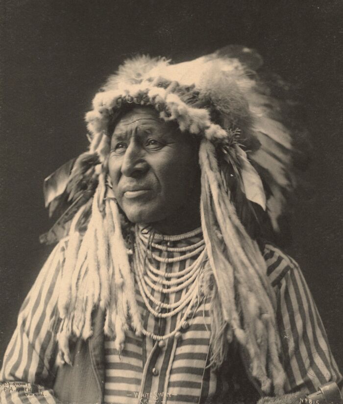 Native American man in traditional headdress and beaded necklaces in a powerful historical portrait from over a century ago.