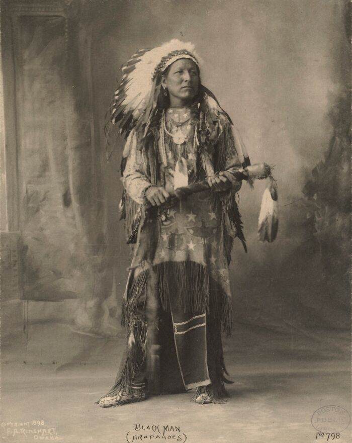 Native American man in traditional attire wearing a feathered headdress and holding a ceremonial staff in a powerful portrait.