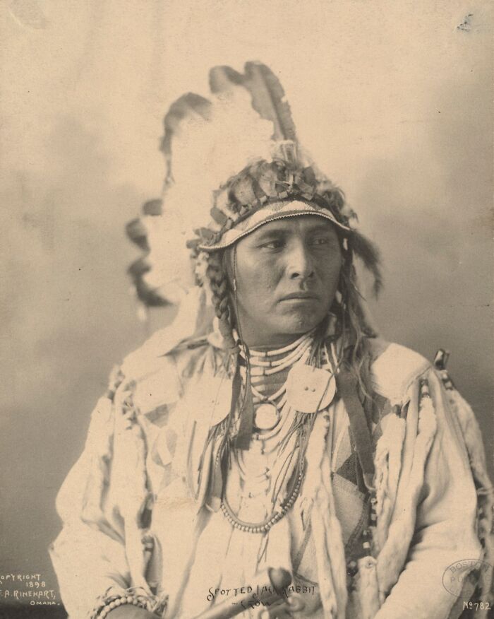 Native American man in traditional headdress and attire, captured in a powerful historic portrait from over a century ago.