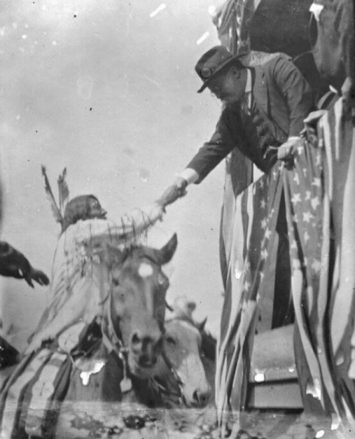 Native American man on horseback shaking hands with a suited man beside an American flag in a historical portrait.