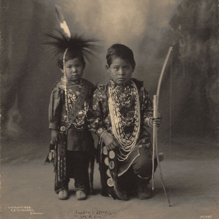 Two Native American boys from a century ago, adorned with traditional clothing, jewelry, and holding a bow in a powerful portrait.