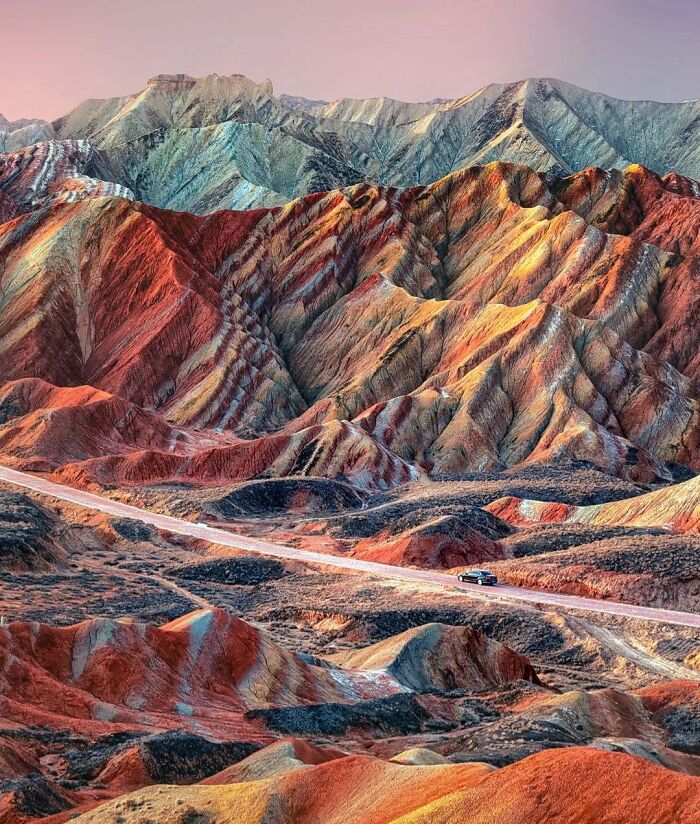 Colorful layered rock formations in a mountainous landscape showcasing rare natural phenomena with a car on a winding road below.