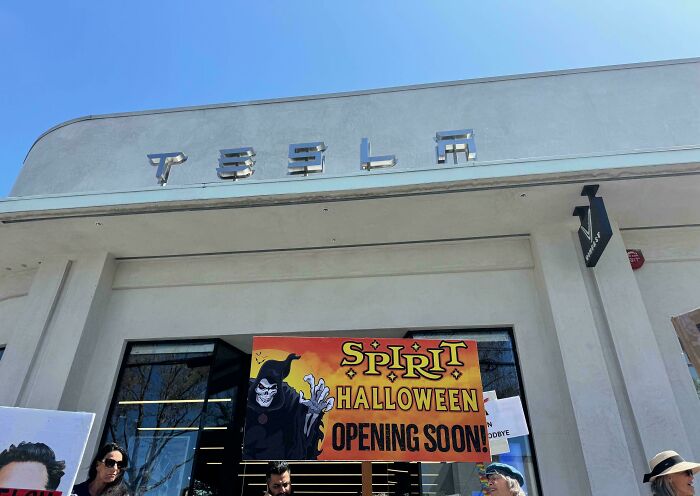 Protesters holding chaotic good signs outside Tesla store with Halloween spirit banner under clear blue sky.