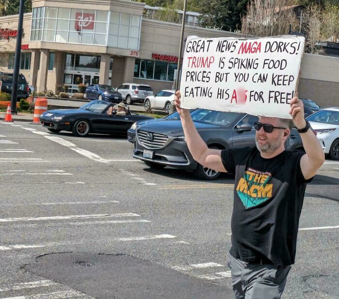 Man holding a chaotic good protest sign in a parking lot with cars and a pharmacy in the background.