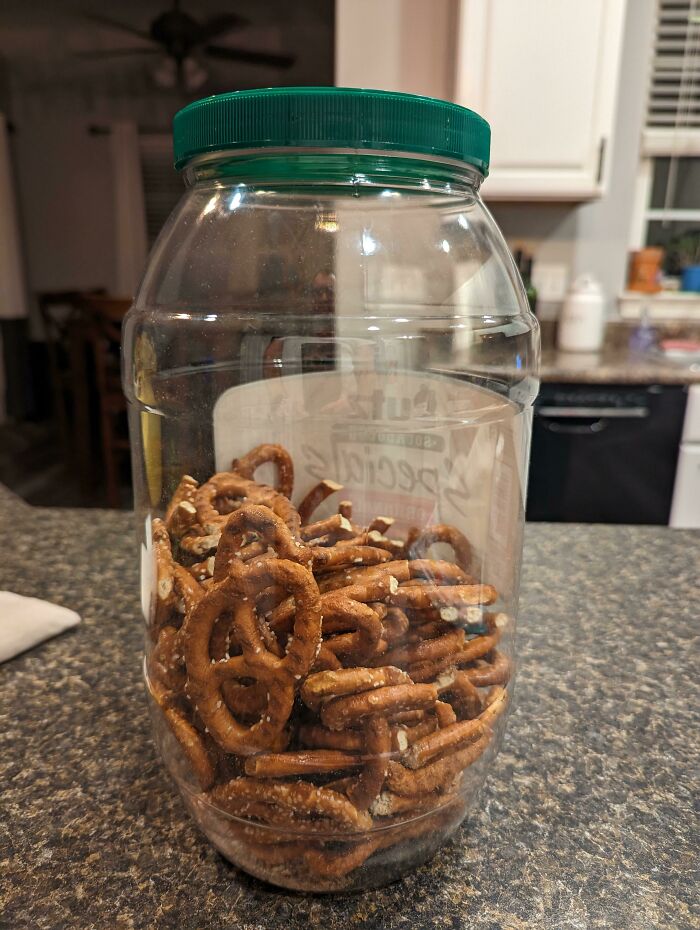 Clear plastic jar with green lid filled with pretzels, illustrating hilariously unlucky parents' snack mishap on kitchen counter.