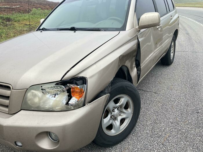 Damaged beige SUV with a broken headlight and dented side panel on a rural road showing unlucky parent car troubles.