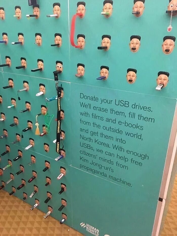 Wall display with USB drives in mouths of Kim Jong-un faces encouraging donations to fight propaganda, highlighting chaotic good acts.