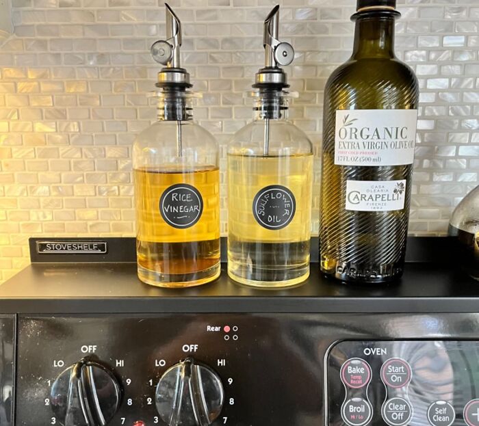 Clear labeled bottles of rice vinegar and sunflower oil with an organic olive oil bottle on a stove shelf for small kitchen solutions.