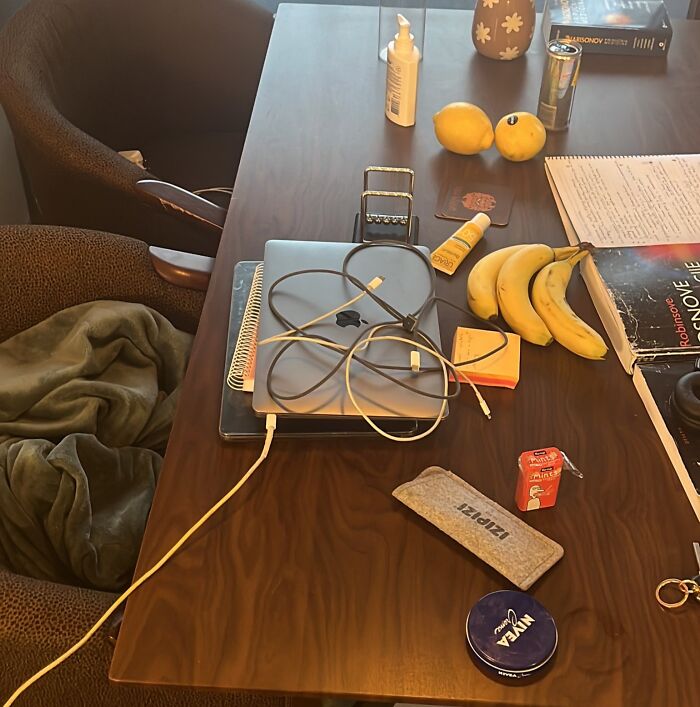 Cluttered home office desk with laptop, charger, snacks, notebook, and personal care items showing a typical work-from-home setup.
