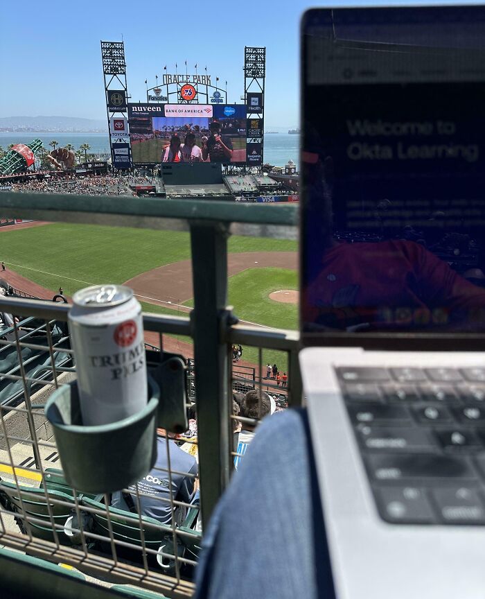 Laptop screen showing remote work setup at a baseball stadium with a drink in a cup holder, illustrating work-from-home memes.