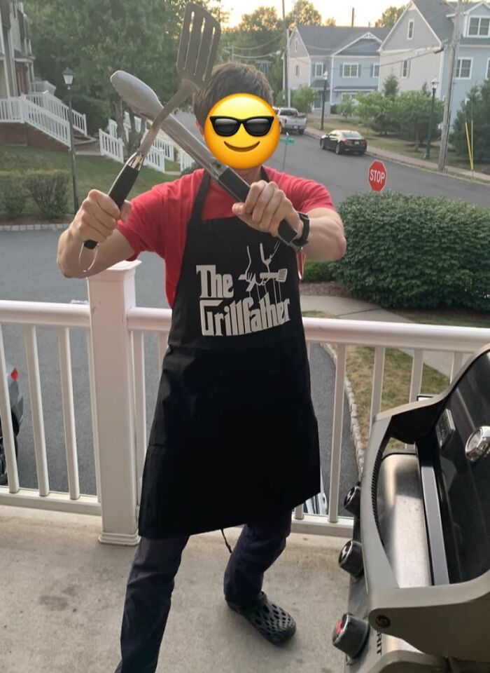 Man wearing The Grillfather apron holding barbecue utensils on porch near grill, perfect Father's Day gifts idea