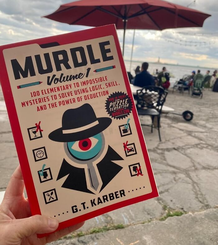 Hand holding Murdle puzzle book outdoors with people and umbrella, perfect Father's Day gifts idea scene.