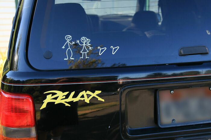 Back of a black car with stick figure family decals and a relaxed vibe, parked near a house on a sunny day.