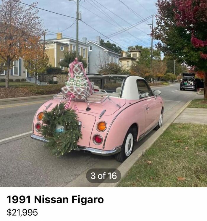Pink 1991 Nissan Figaro parked on a street decorated with a holiday wreath and wrapped gifts for sale.
