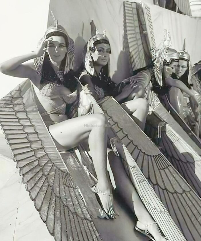 Women dressed in ancient history costumes with winged accessories sitting in a row, showcasing historical fashion designs.