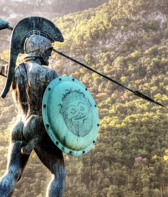 Ancient history warrior statue with spear and shield featuring a mythological face, set against a scenic forested mountain background.