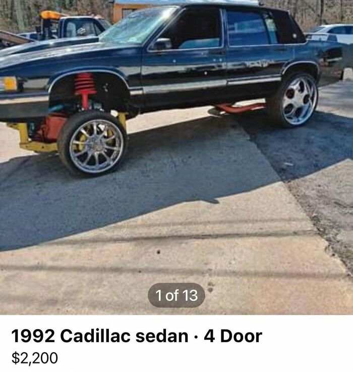 Black 1992 Cadillac sedan with large chrome wheels and raised suspension in a chaotic car sale setting.